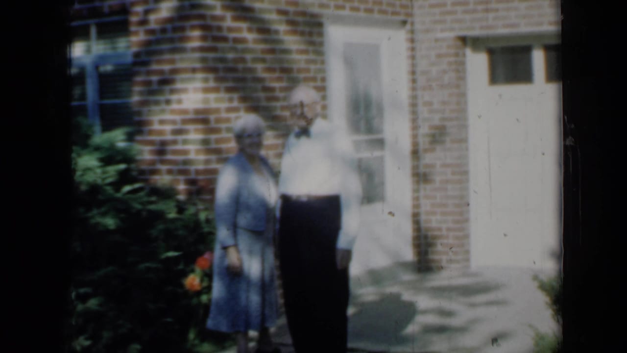 1958 Minnesota home-movie frame of an elderly couple standing outside a brick residence in tree shade near a garage and window.