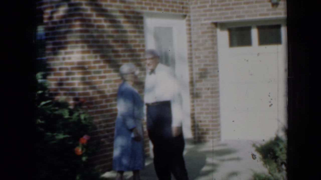 1958 Minnesota home-movie frame showing the couple turned toward each other outside the brick home, with shrubs and a garage door visible.