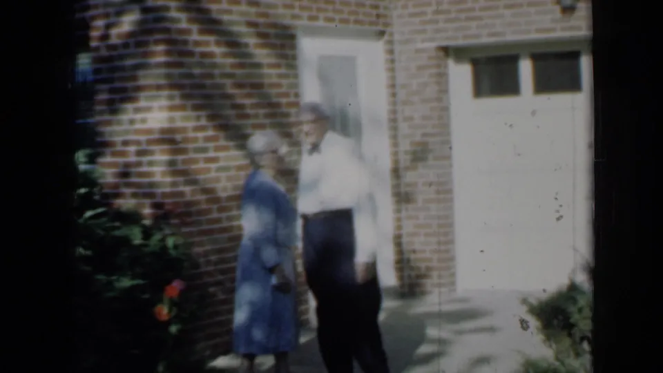 1958 Minnesota home-movie frame showing the couple turned toward each other outside the brick home, with shrubs and a garage door visible.