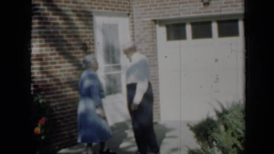 1958 Minnesota home-movie frame of the couple standing apart in shade beside a brick wall, window, walkway, and white garage door.