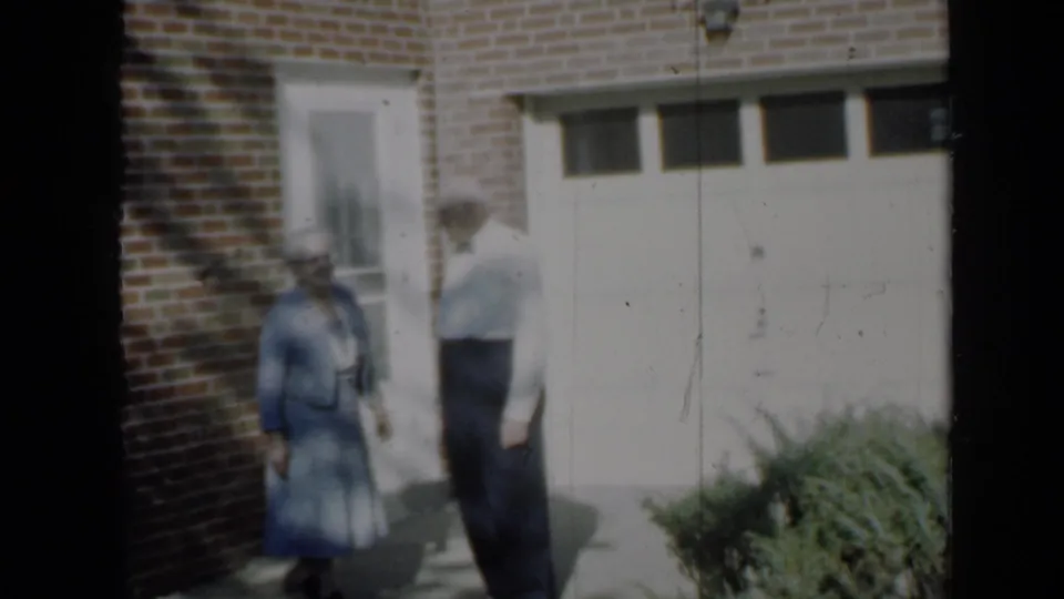 1958 Minnesota home-movie frame showing the elderly couple in front of a brick residence, with dappled shade across the wall and ground.
