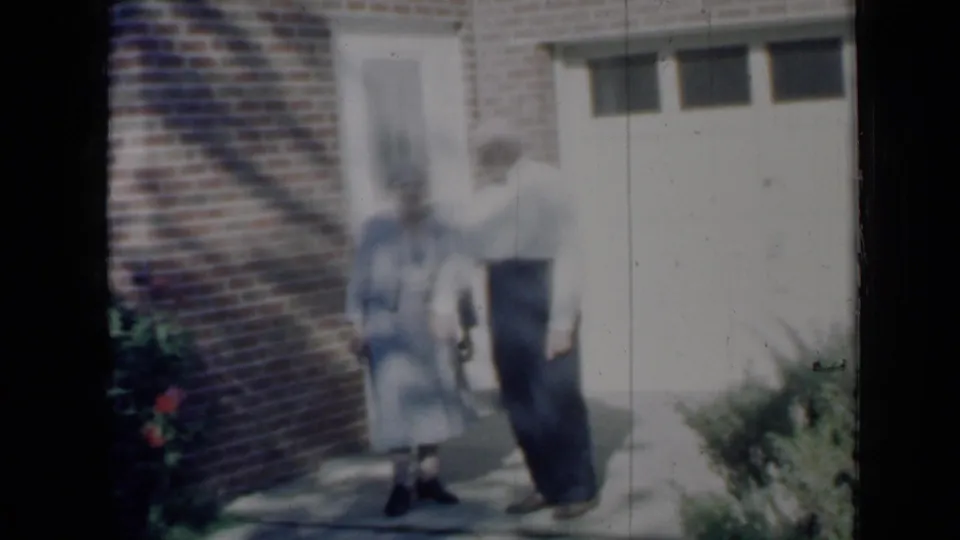 Elderly couple outside a brick Minnesota house in 1958 home-movie footage, standing near a doorway before approaching a car.