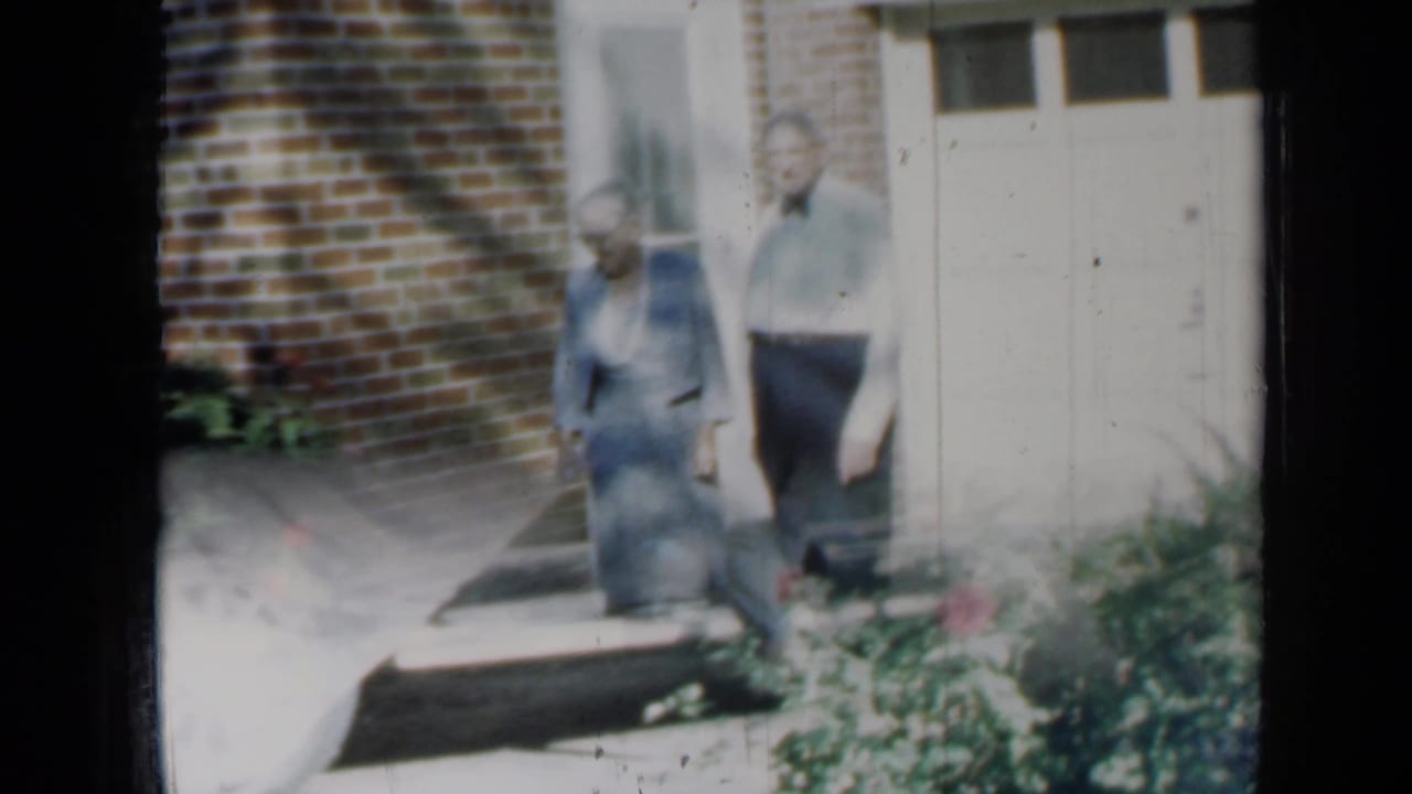 Elderly couple beside a brick house with garden plants in front.