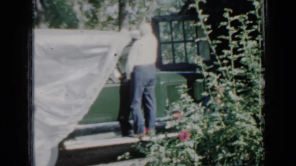 An older man pulls back a white cover from a green car beside a brick Minnesota house in 1958 home-movie footage.