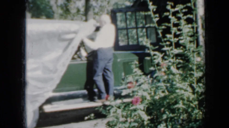 The green car is partly uncovered as the older man stands beside it near a brick house and garden in Minnesota footage from 1958.