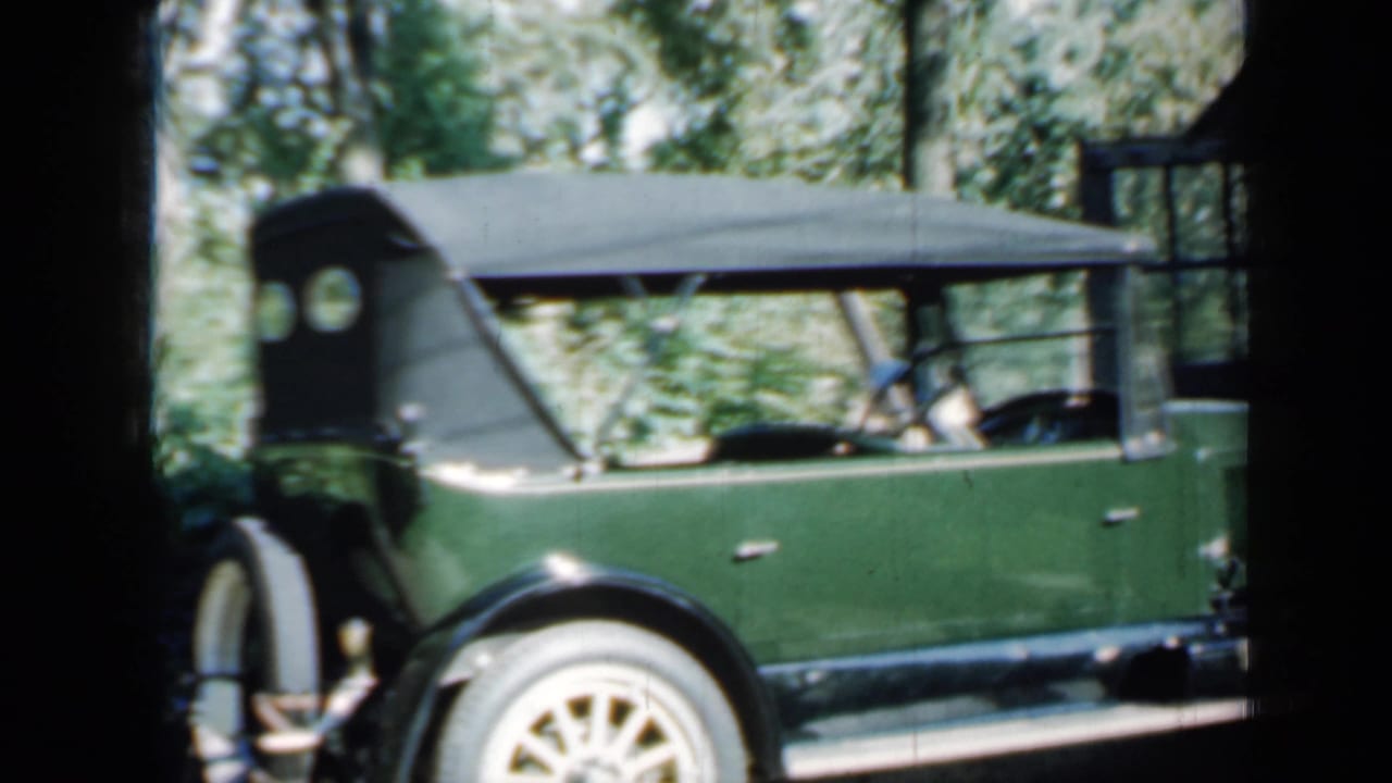 Green open-top vintage car parked beside trees and a rustic building.