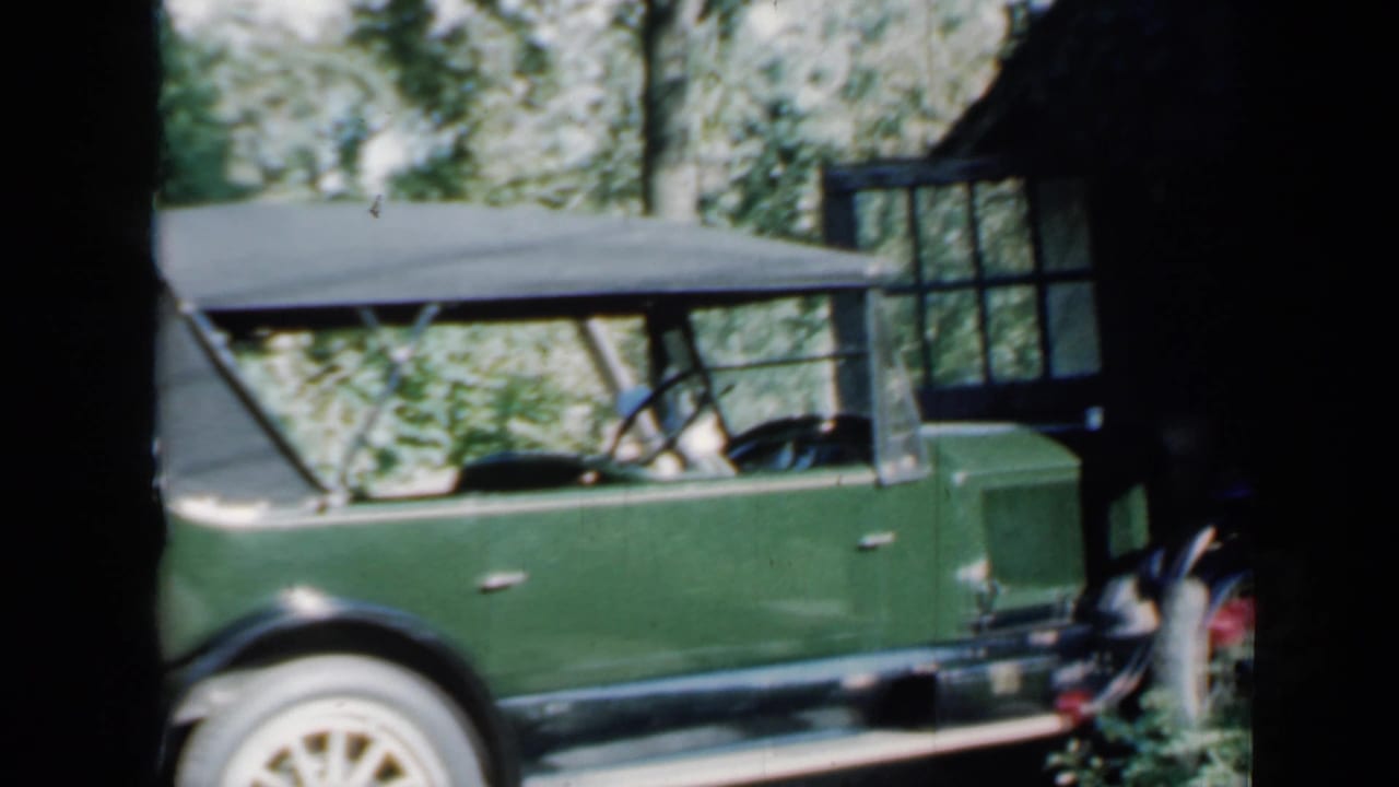 Side view of the green classic car with black soft top, chrome details, and wooded background in archival home-movie footage.