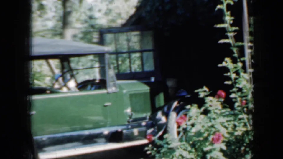 Rear section of a green classic car near garden flowers, trees, and a dark rustic structure in the 1958 Minnesota home-movie clip.