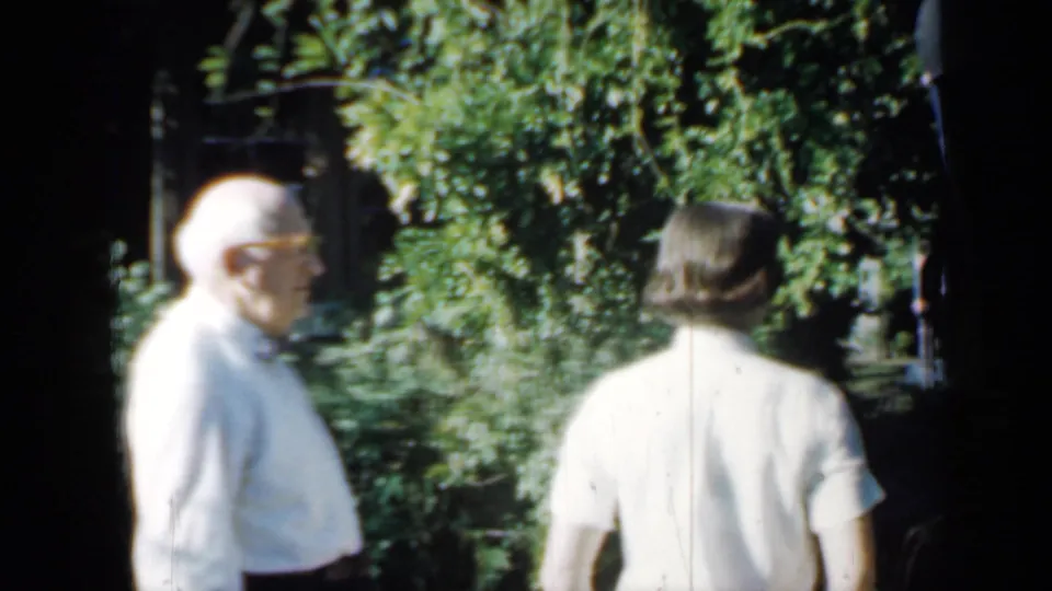Two people stand outdoors among greenery in the Minnesota home-movie scene, with faces and bodies partly visible in bright sun.