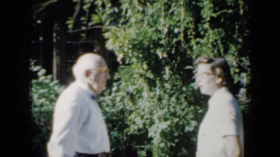 In 1958 Minnesota home-movie footage, an older man in a white shirt and bow tie faces a younger woman in a garden setting.