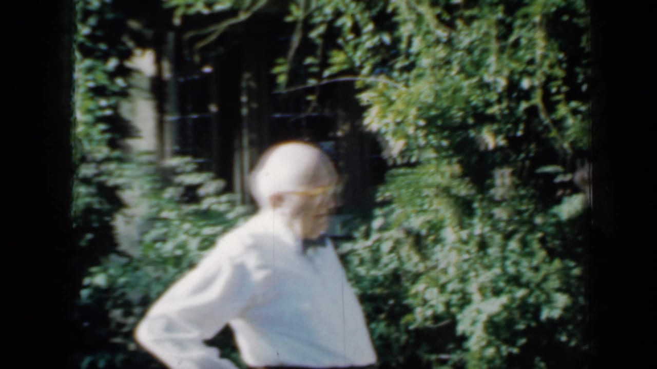 Older man in a white shirt and bow tie stands in a leafy Minnesota garden area, filmed on 8mm in 1958.