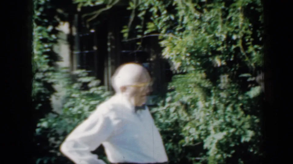 Older man in a white shirt and bow tie stands in a leafy Minnesota garden area, filmed on 8mm in 1958.