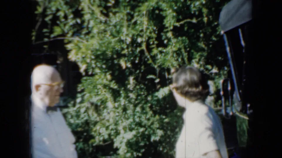 The older man and younger woman appear partly framed by the handheld camera near dense foliage in the 1958 Minnesota garden scene.