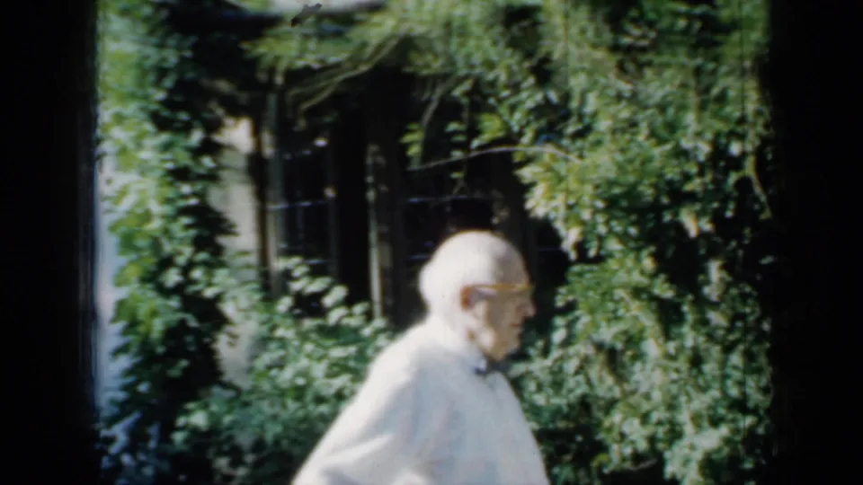 Older man in profile, wearing a white shirt and bow tie, stands by sunlit greenery in the supported 1958 Minnesota home-movie clip.