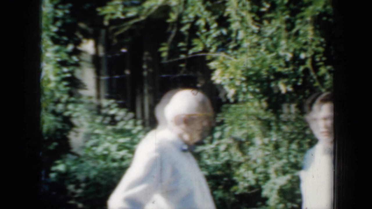 In 1958 Minnesota home-movie footage, an elderly man stands near dense garden plants while another person is partly visible beside a brick house.