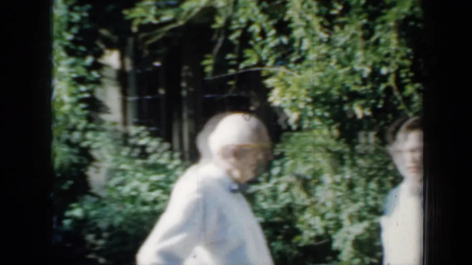 In 1958 Minnesota home-movie footage, an elderly man stands near dense garden plants while another person is partly visible beside a brick house.