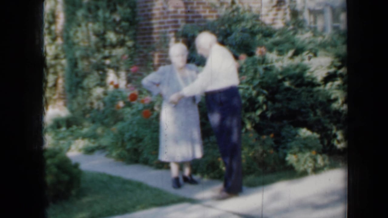 An elderly couple in 1958 Minnesota footage stands on a narrow garden path near roses, shrubs, and a brick residential wall.