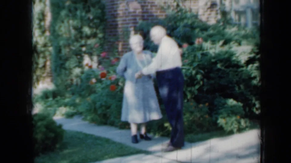The elderly couple appears to pose on a paved garden walk in Minnesota, with rose bushes, shrubs, lawn, and brickwork around them.