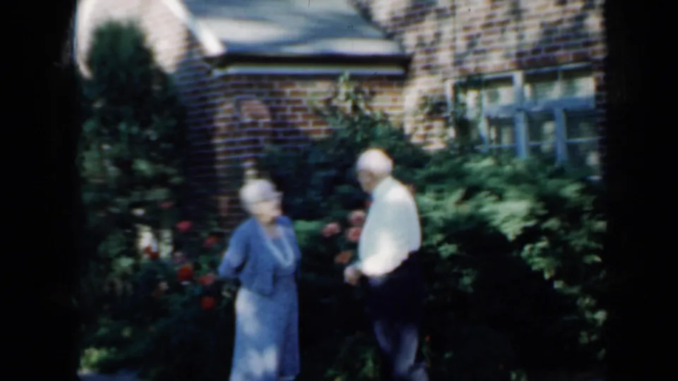 The couple stands near rose bushes and shrubs in front of a brick house, with windows and strong sunlight visible in the frame.