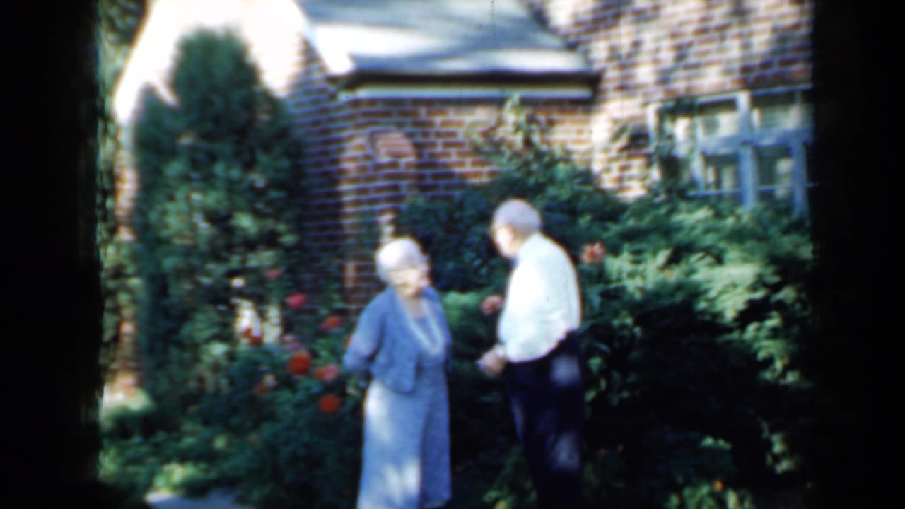 Elderly couple standing in a sunlit garden outside a brick house.