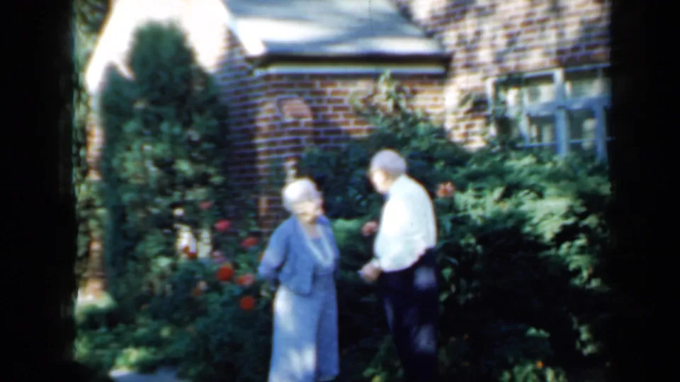 Elderly couple in 1958 Minnesota home-movie footage standing beside flowers and shrubs outside a brick residence.