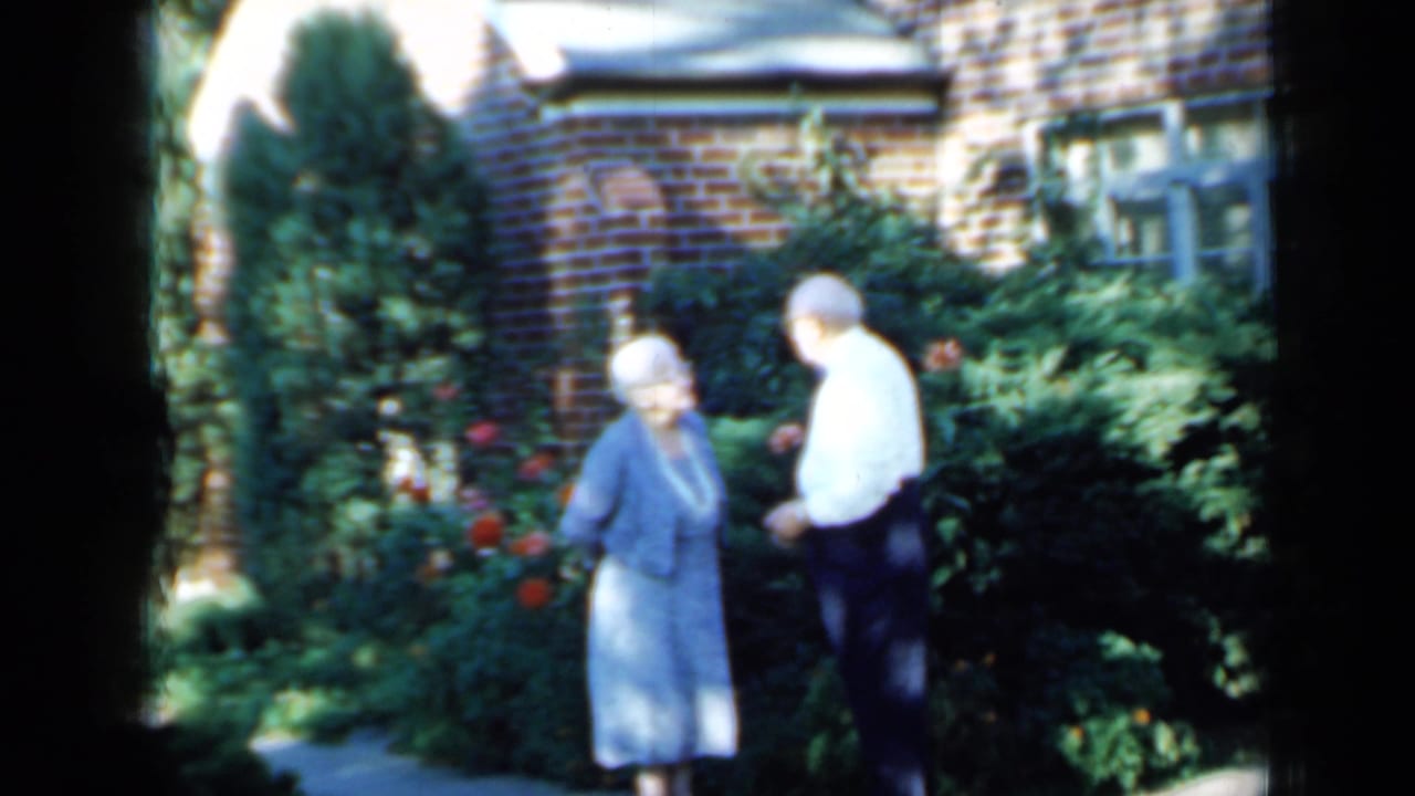 Elderly woman in blue and man in a white shirt appear to converse in a Minnesota residential garden, 1958.