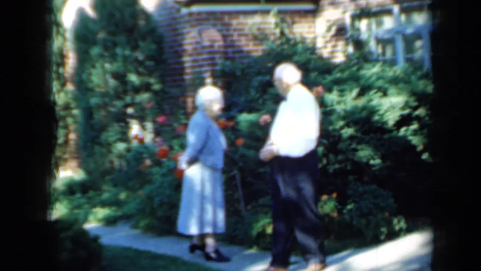 The couple stands closer together on a paved path amid shrubs and flowers outside a brick Minnesota residence in 1958 footage.