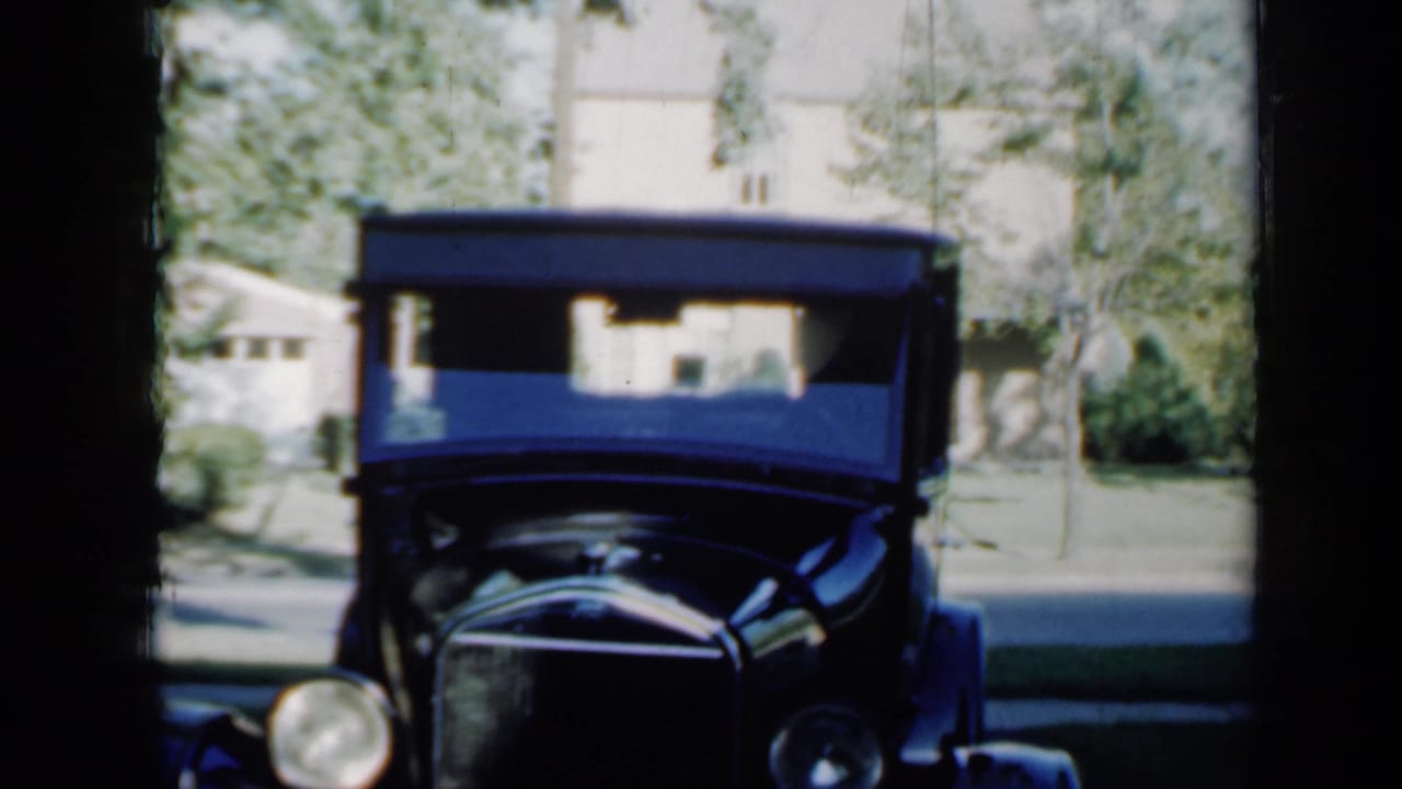 1958 Minnesota home-movie frame showing an antique black car parked in a driveway, viewed through deep shade with a house and trees behind.