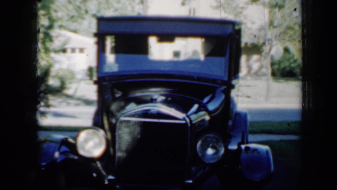 1958 Minnesota frame with the antique car more centered, its tall windshield, rounded headlights, grille, driveway, and background house visible.