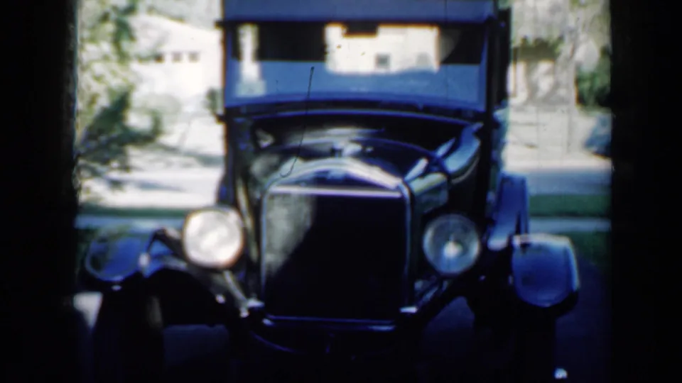 1958 Minnesota home-movie frame showing the antique car closer in view, with its headlights, hood, grille, driveway, trees, and house visible.