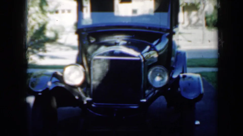 Final 1958 Minnesota frame showing the antique car from the front, with deep shadow in the foreground and a sunlit suburban driveway behind.