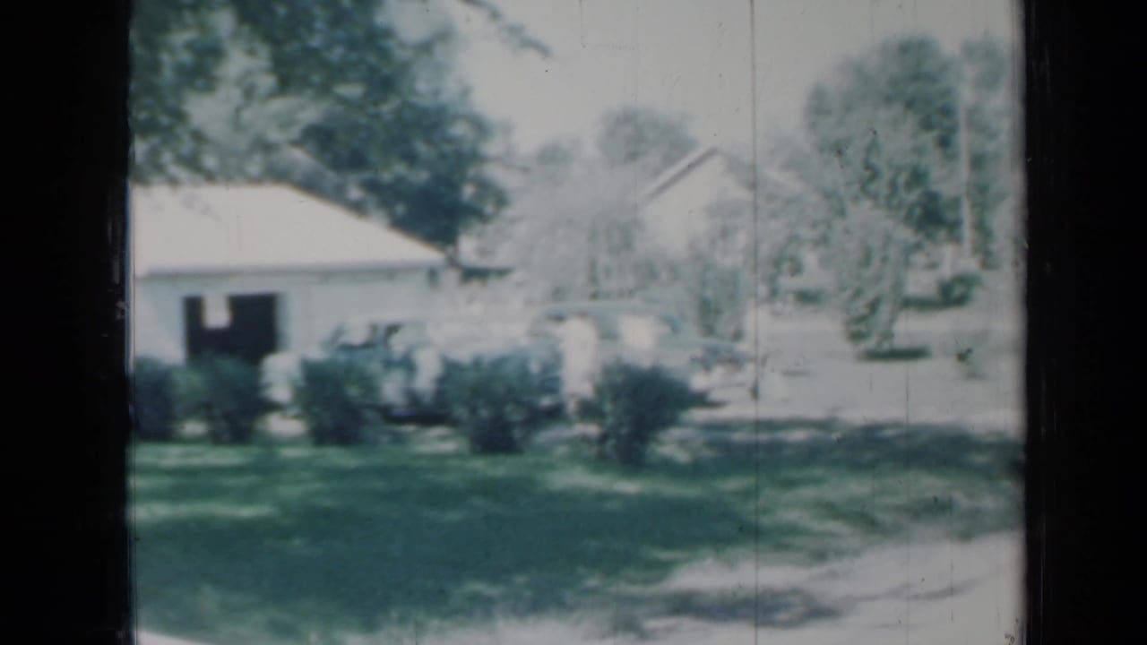 1958 Minnesota 8mm frame showing a white house, garage area, green lawn, bushes, and trees in soft home-movie focus.