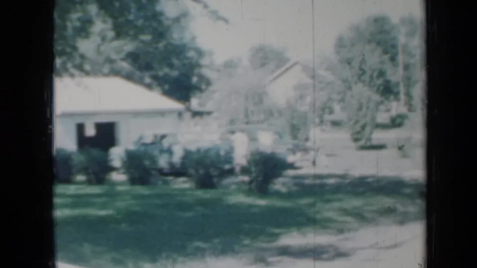 1958 Minnesota 8mm frame showing a white house, garage area, green lawn, bushes, and trees in soft home-movie focus.