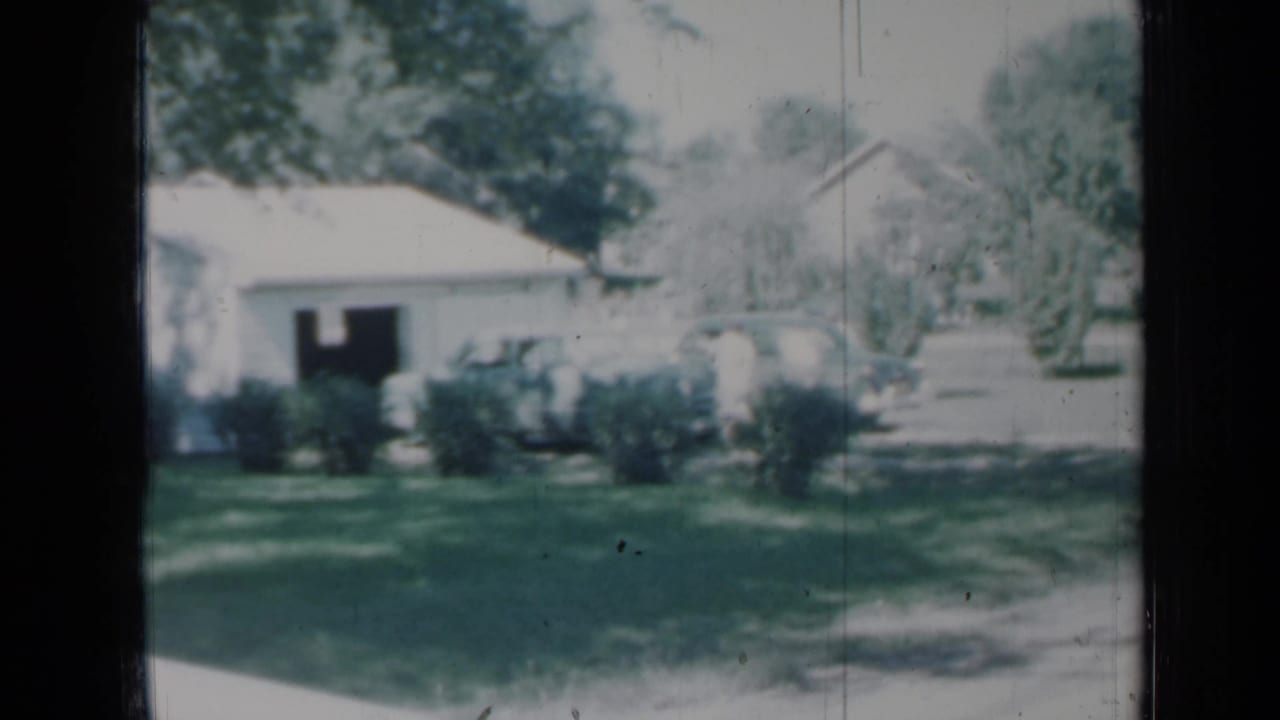 A soft 1958 Minnesota home-movie view of a white house with shrubs, lawn, trees, and a driveway area.
