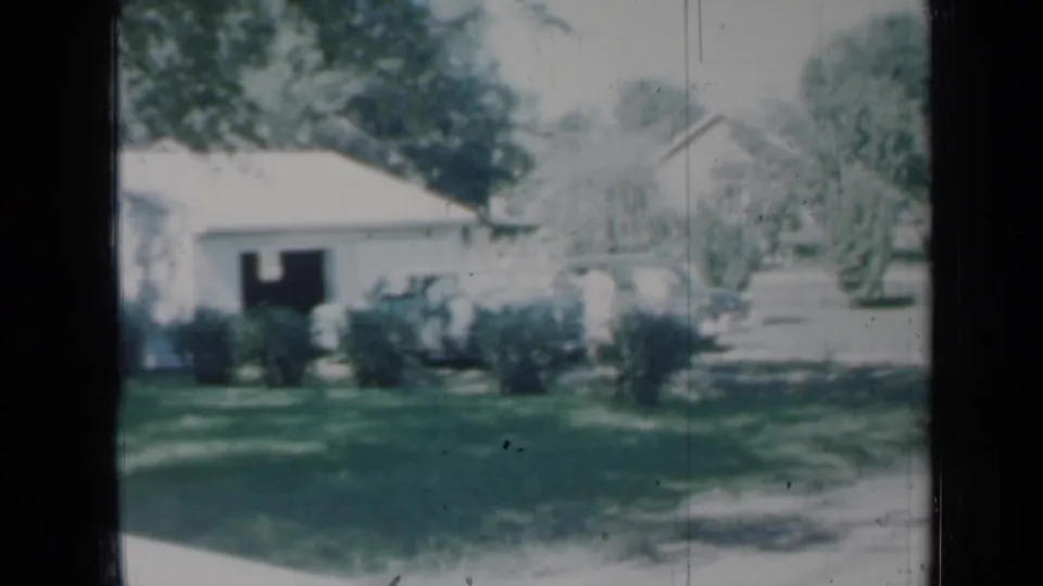 A soft 1958 Minnesota home-movie view of a white house with shrubs, lawn, trees, and a driveway area.