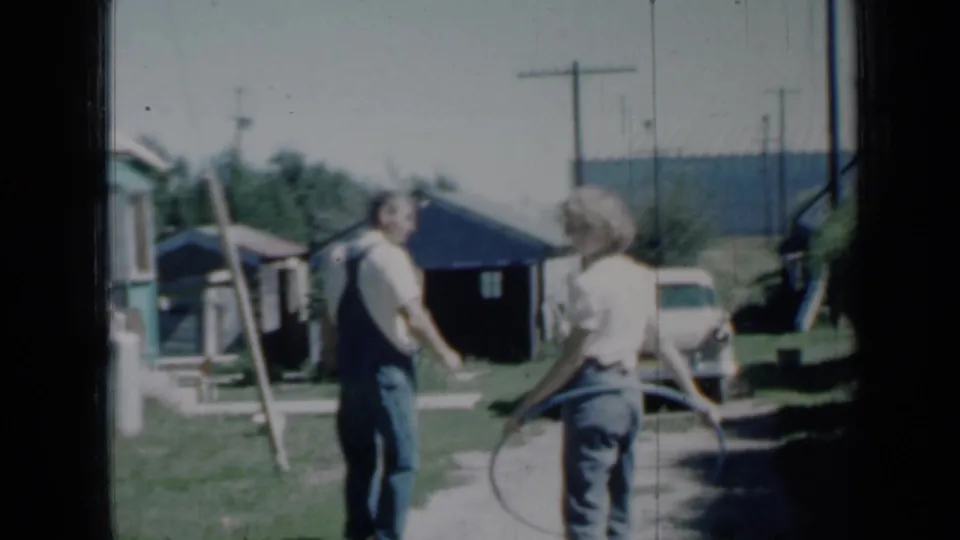 In a 1958 Minnesota yard, a man in overalls and a girl with a hoop walk near a driveway, shed, car, and utility poles.