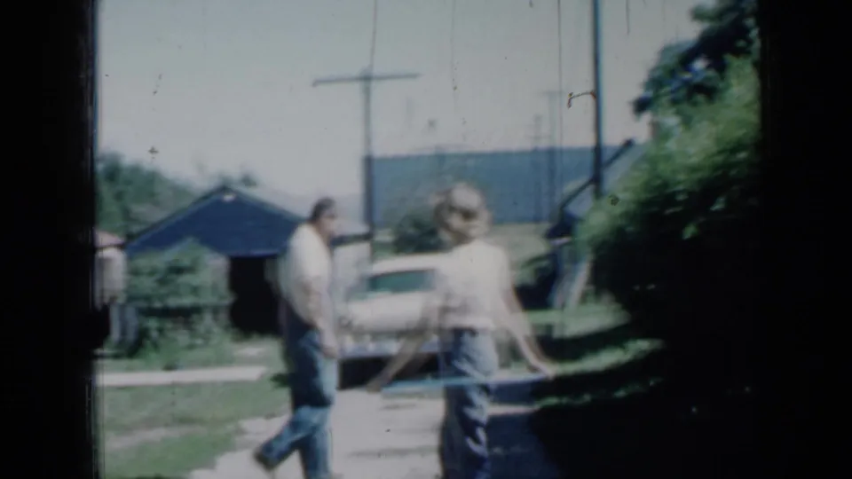 The 1958 Minnesota home-movie frame shows a girl holding a hoop and a man walking past a driveway with a shed and parked car.