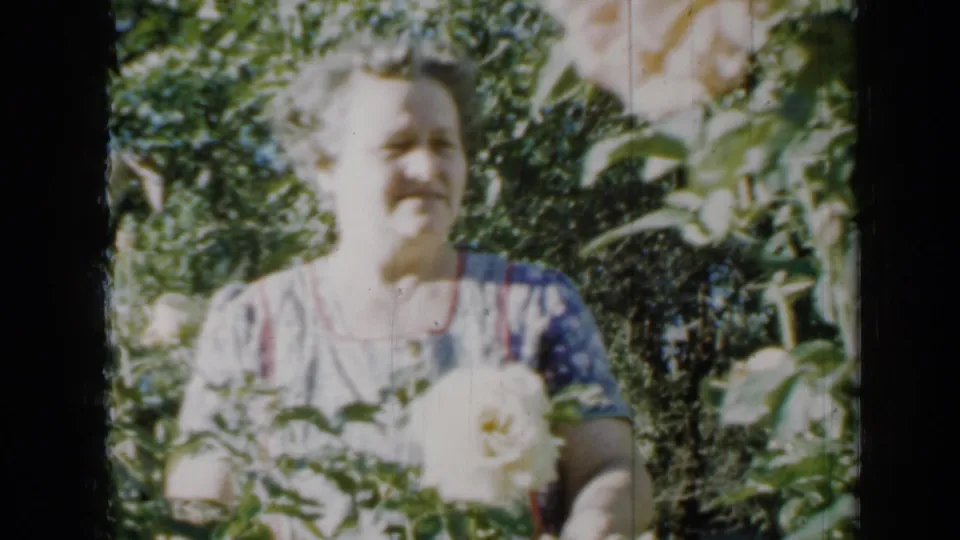 Adult woman in a patterned dress stands among garden plants in 1958 Minnesota, holding a white rose.