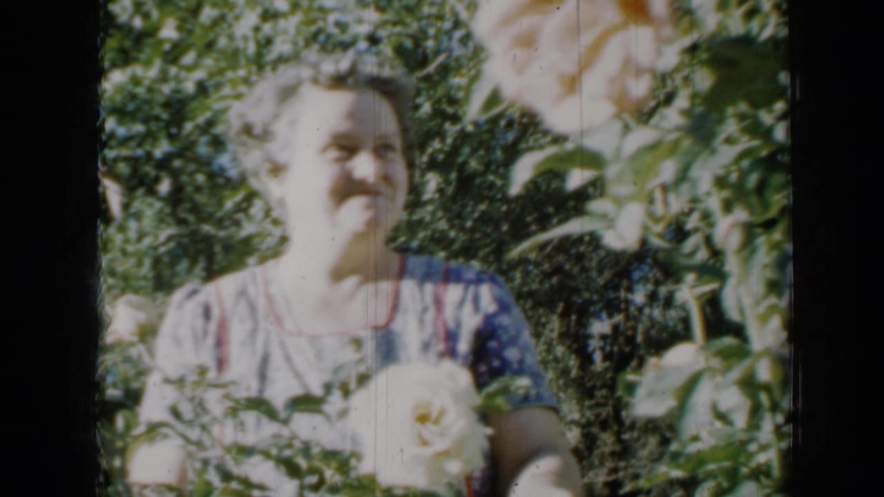 In 1958 Minnesota home-movie footage, a woman in a patterned dress smiles near pale roses in a sunlit garden.