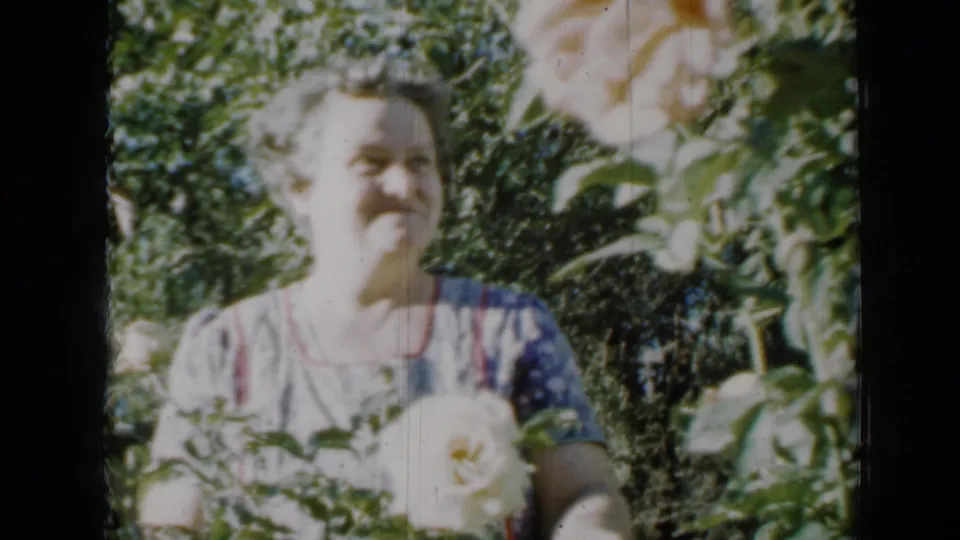In 1958 Minnesota home-movie footage, a woman in a patterned dress smiles near pale roses in a sunlit garden.