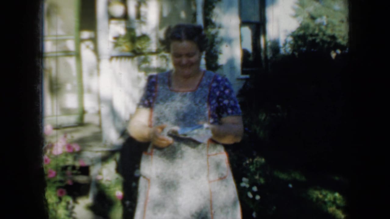 Woman in apron standing on a sunlit porch near flowers and shrubs.