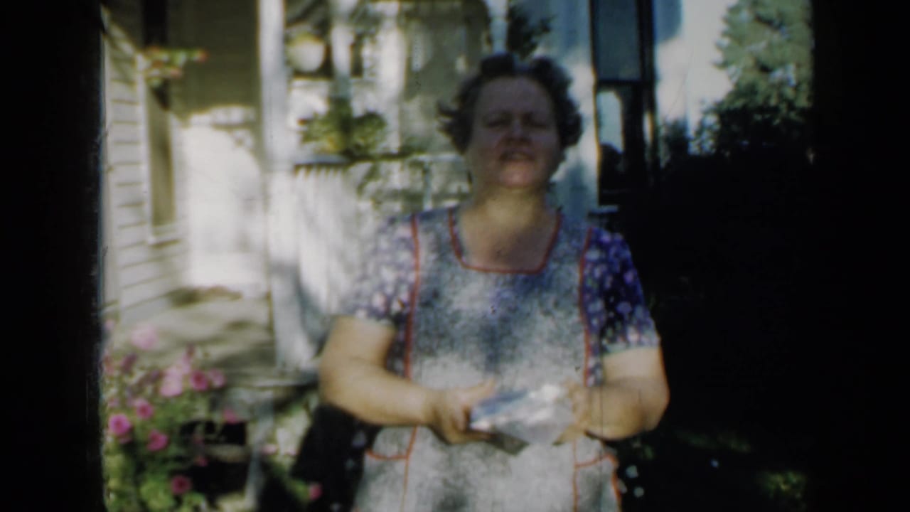 A woman in a patterned dress and apron faces the camera on a Minnesota porch, holding a small object amid bright sun and shade.