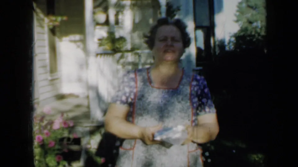 A woman in a patterned dress and apron faces the camera on a Minnesota porch, holding a small object amid bright sun and shade.
