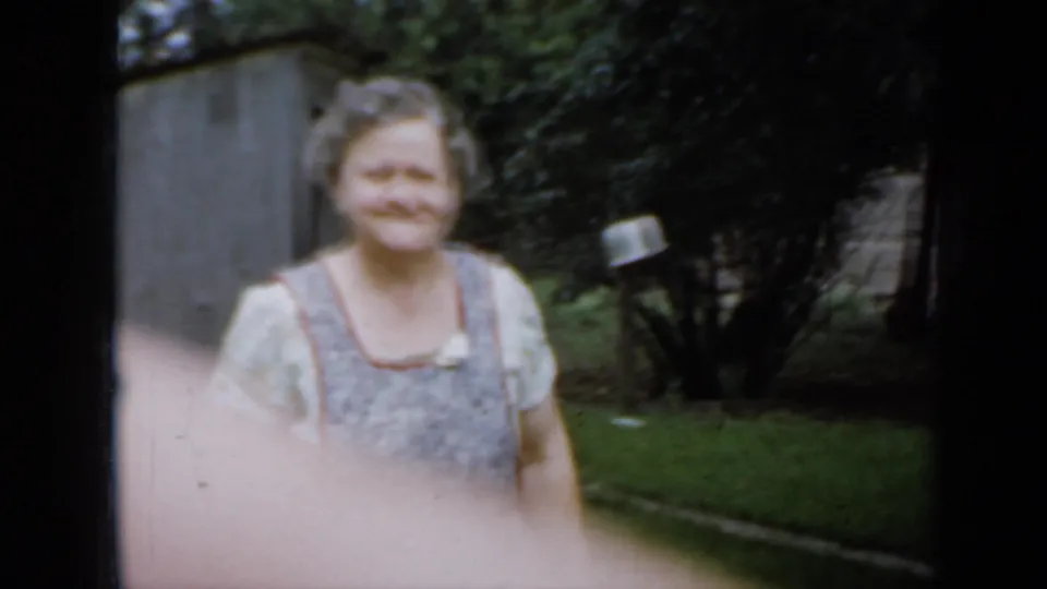 Older woman in a light dress and patterned apron smiles toward the camera in a Minnesota garden setting, 1958.
