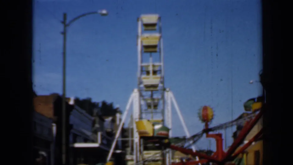 1958 Minnesota carnival view with a white Ferris wheel, red spinning ride structure, colored gondolas, and storefront signs.