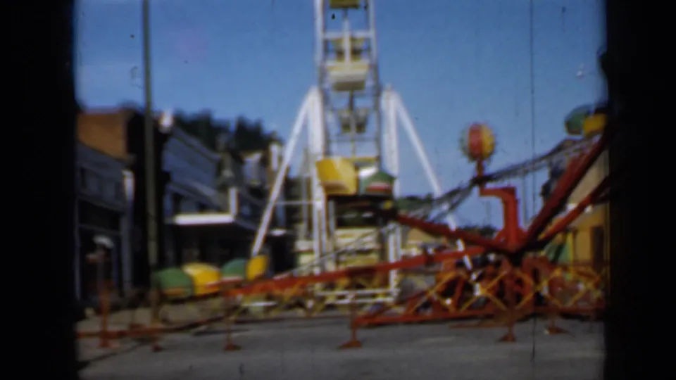 1958 Minnesota 8mm frame showing a red spinning ride with colored cars, a Ferris wheel, storefronts, and clear sky.