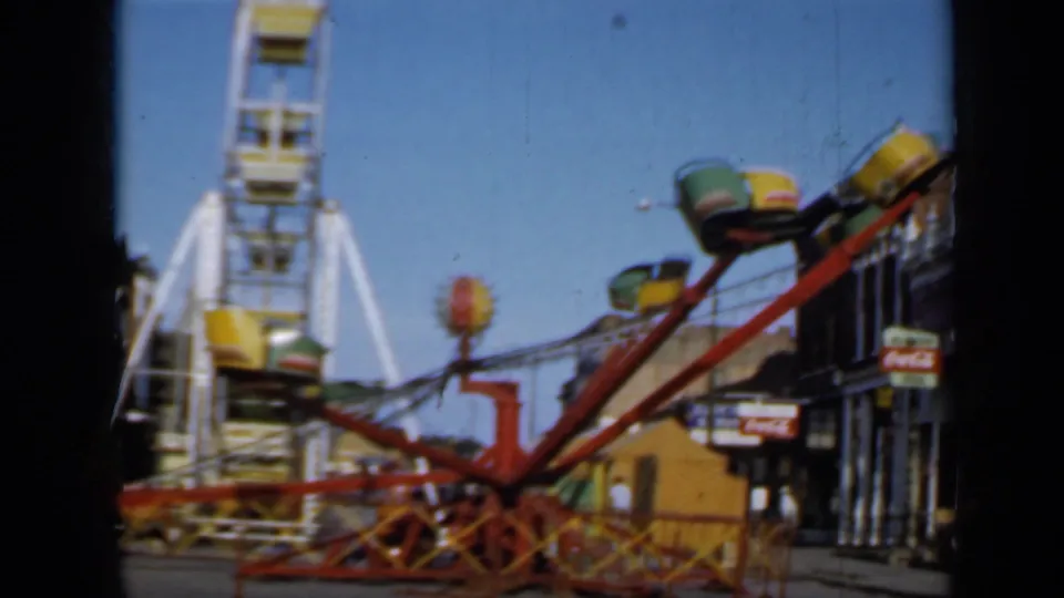 1958 Minnesota carnival frame with a red ride, colored gondolas, Ferris wheel, storefronts, and a visible Coca-Cola sign.
