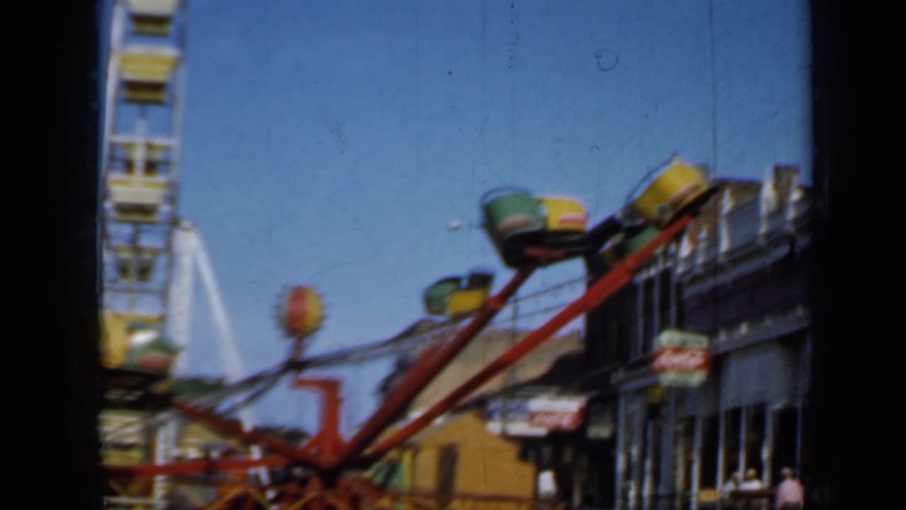 Red spinning carnival ride and Ferris wheel beside small-town storefronts.