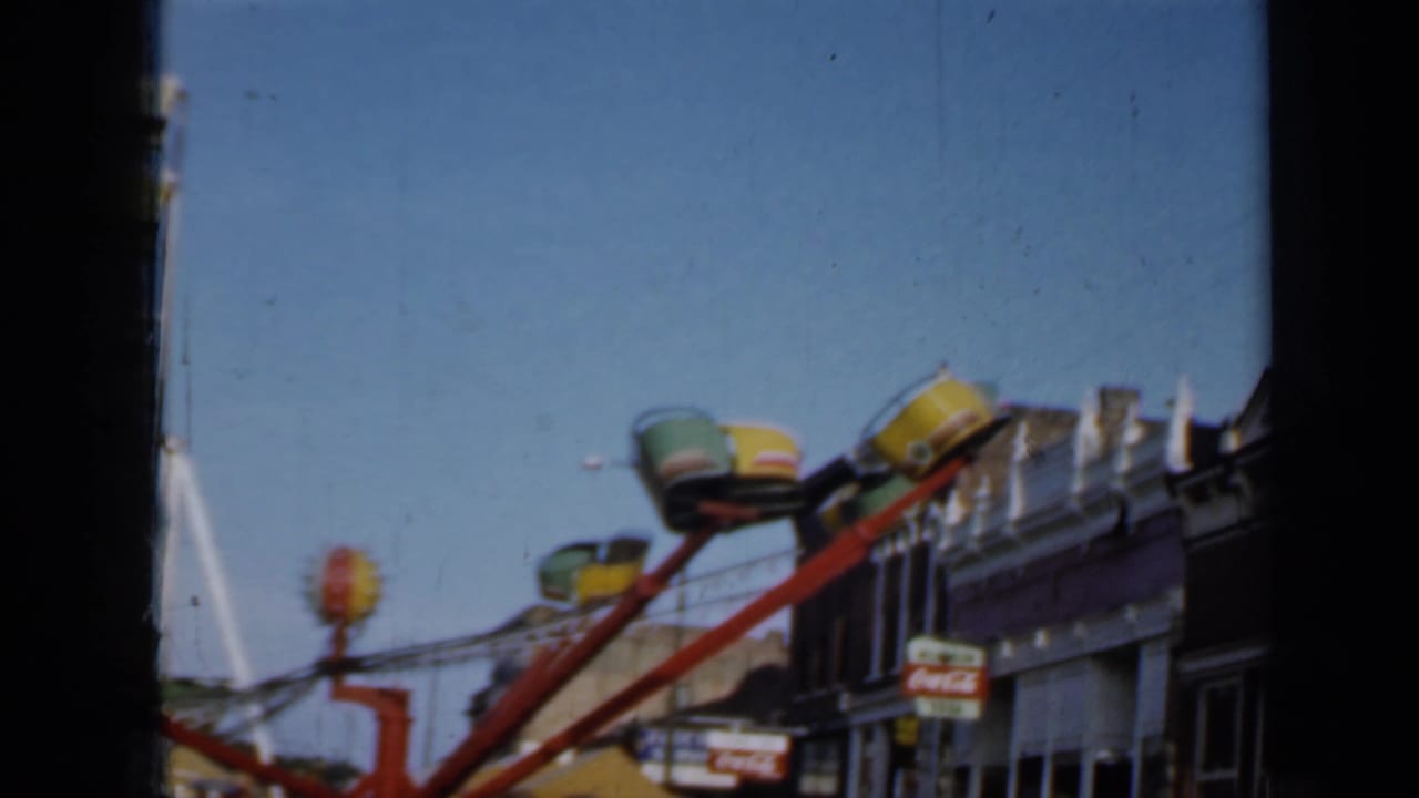 1958 Minnesota fairground view with empty ride seats, a red ride arm, storefront facades, and a visible Coca-Cola sign.