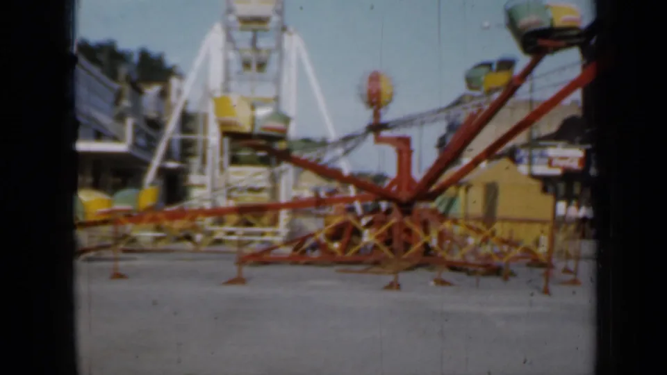 1958 Minnesota fairground frame with an idle red ride, empty bucket seats, a white Ferris wheel, and storefronts along the midway.